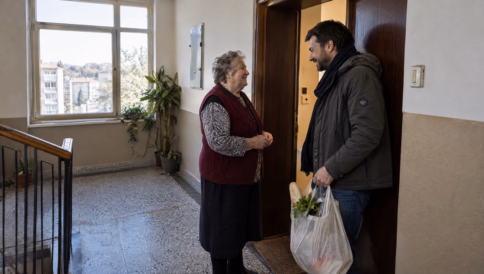 Starija komšinica i komšija sa kesom iz prodavnice razgovaraju na odmorištu stambene zgrade.