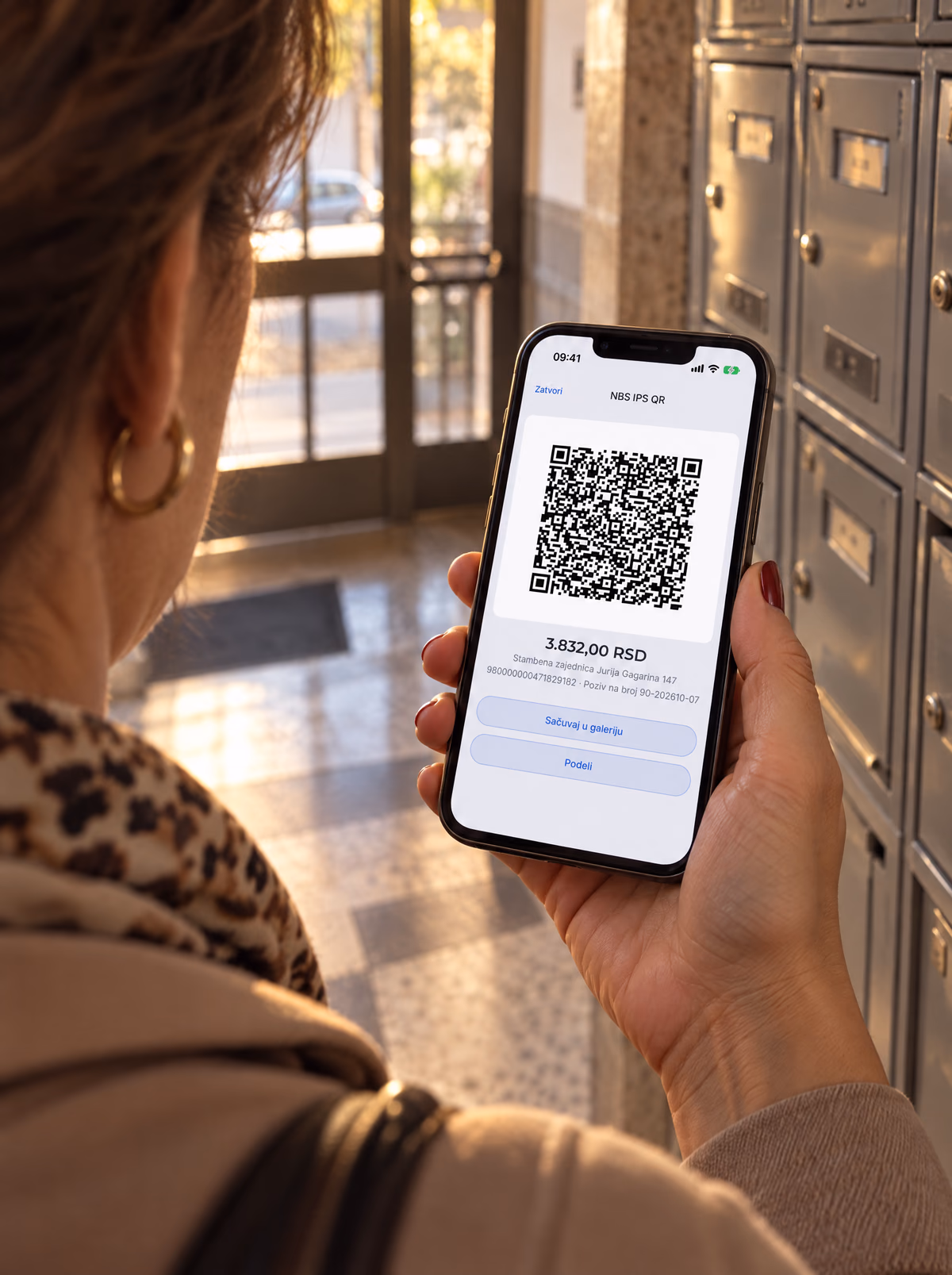 Žena u ulazu stambene zgrade, pored poštanskih sandučića, drži telefon. Na ekranu je IPS QR kod računa u aplikaciji esz.rs, sa primerom podataka.